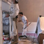 Two painters working indoors, one standing on a stool.