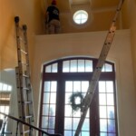 Two workers on ladders installing or repairing near a high window inside a building.
