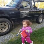 Toddler in pink outfit near a black pickup truck on a gravel path.