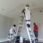 Two workers painting a ceiling while a man observes.