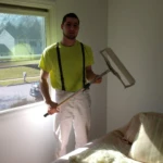 Man in workwear holding a floor cleaning tool in a sunlit room.