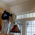 Painter on a ladder painting a wall near windows.