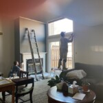 Two people working on home renovation near large windows and a ladder.